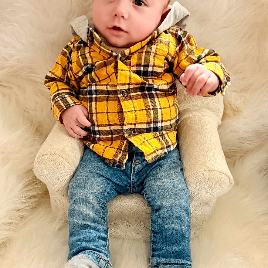 Roman is registered to the contest to win money with this photo: baby, child, plaid_shirt, yellow_shirt, blue_jeans, socks, small_chair, cushion, fluffy_background, indoor, portrait, seated, infant, cute, looking_at_camera, casual_clothing, soft_texture, comfortable, young_child, curious