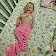 Hope is registered to the contest to win money with this photo: baby, crib, mermaid_costume, pink, smiling, infant, bottle, plush_toy, blanket, patterned_sheet, cute, happy, sleepwear, child, indoors, resting, decor, soft_toys, knitted, playful