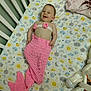 baby, crib, mermaid_costume, pink, smiling, infant, bottle, plush_toy, blanket, patterned_sheet, cute, happy, sleepwear, child, indoors, resting, decor, soft_toys, knitted, playful