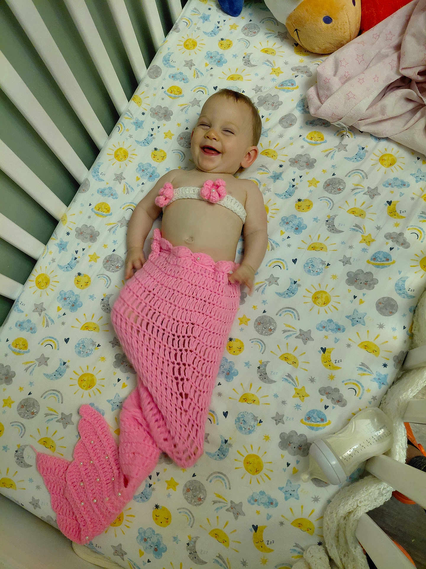 Hope is registered to the contest to win money with this photo: baby, crib, mermaid_costume, pink, smiling, infant, bottle, plush_toy, blanket, patterned_sheet, cute, happy, sleepwear, child, indoors, resting, decor, soft_toys, knitted, playful