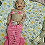 baby, smiling, mermaid_costume, pink, crib, baby_bottle, blanket, patterned_sheet, cute, infant, lying_down, happy, child, clothing, toddler, hand, face, playful, indoors, nursery