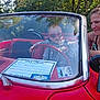 adult, baby, car, car_show, child, clothing, convertible, fun, heart_shaped, license_sticker, outdoor, person, red_car, smile, steering_wheel, sunglasses, sunlight, trees, vintage_car, woman