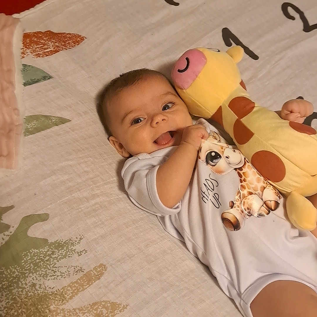 Zahara is registered to the contest to win money with this photo: baby, blanket, child, clothing, comfort, cute, face, giraffe, hand, happy, head, indoors, infant, lying_down, person, playful, plush_toy, smiling, soft_toy, toy