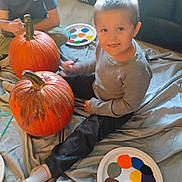 Leonidas is registered to the contest to win money with this photo: child, pumpkin, painting, paint_plate, paintbrush, smile, indoor, blanket, socks, cap, art_activity, craft, floor, pair_of_shoes, autumn, family_activity, portrait, toddler, clothing, playful