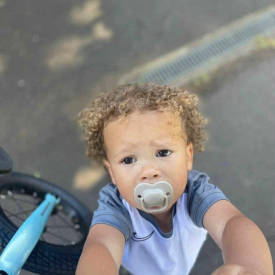 Reyes is registered to the contest to win money with this photo: bicycle, blue_shirt, child, closeup, curious, curly_hair, cute, daylight, expression, looking_up, outdoor, pacifier, pavement, person, reaching, short_sleeves, toddler, wheel, white_shirt, young_child