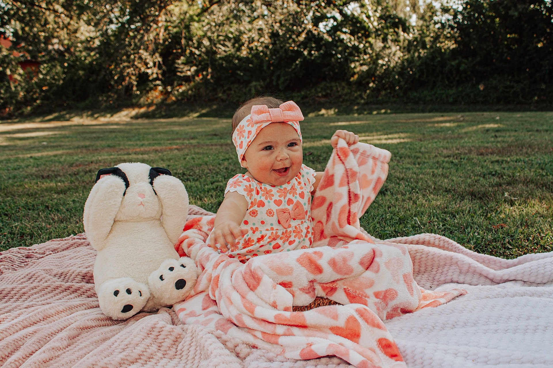 Ivory is registered to the contest to win money with this photo: baby, blanket, child, cute, dog_toy, floral_clothing, grass, happy, headband, heart_pattern, nature, outdoor, pink, playful, plush_toy, portrait, sitting, smiling, sunlight, toy