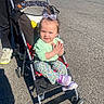 toddler, stroller, child, outdoor, parking_lot, clothing, bow, happy, person, sneakers, sunlight, pavement, baby, casual_wear, wheelchair, seat, toy, playful, daytime, smiling
