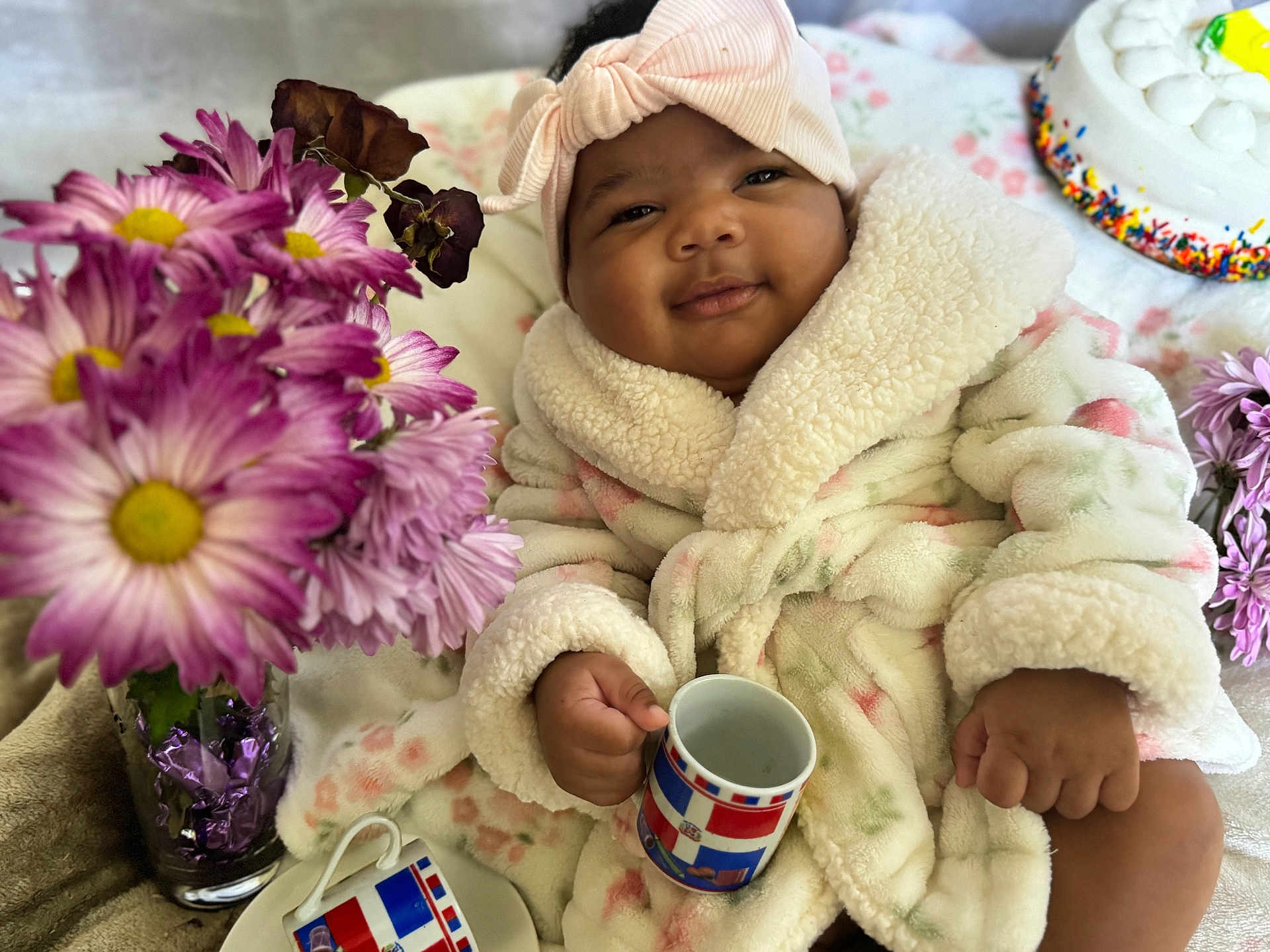 Jameline joined the competition — help win amazing prizes! baby, child, smiling, robe, headband, bow, mug, flowers, purple_flowers, cake, table, indoor, soft_texture, cute, portrait, happy, infant, warm_clothing, decor, celebration