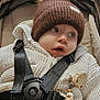 baby, brown_hat, child, closeup, comfortable, curious, cute, expression, face, indoors, infant, person, pom_pom, safety_harness, seatbelt, soft_lighting, stroller, warm_clothing, white_jacket, zipper
