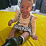 boots, bow, child, curtains, dress, fan, floor, footwear, furniture, glasses, hand, happy, headband, indoor, leopard_print, person, sitting, smiling, toddler, yellow_blanket