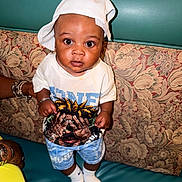 Tymonte is registered to the contest to win money with this photo: baby, child, infant, portrait, standing, couch, sofa, cushion, white_hat, white_shirt, shorts, socks, toy, wide_eyes, curious, indoors, seated_area, floral_pattern, teal_background, photography