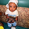 baby, child, infant, portrait, standing, couch, sofa, cushion, white_hat, white_shirt, shorts, socks, toy, wide_eyes, curious, indoors, seated_area, floral_pattern, teal_background, photography