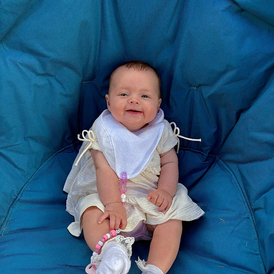 Poppy joined the competition — help win amazing prizes! baby, baby_accessory, bib, blue_cushion, casual, child, comfortable, cute, happy, indoor, infant, lace, pacifier_clip, person, portrait, seated, smiling, socks, white_clothing, young_child
