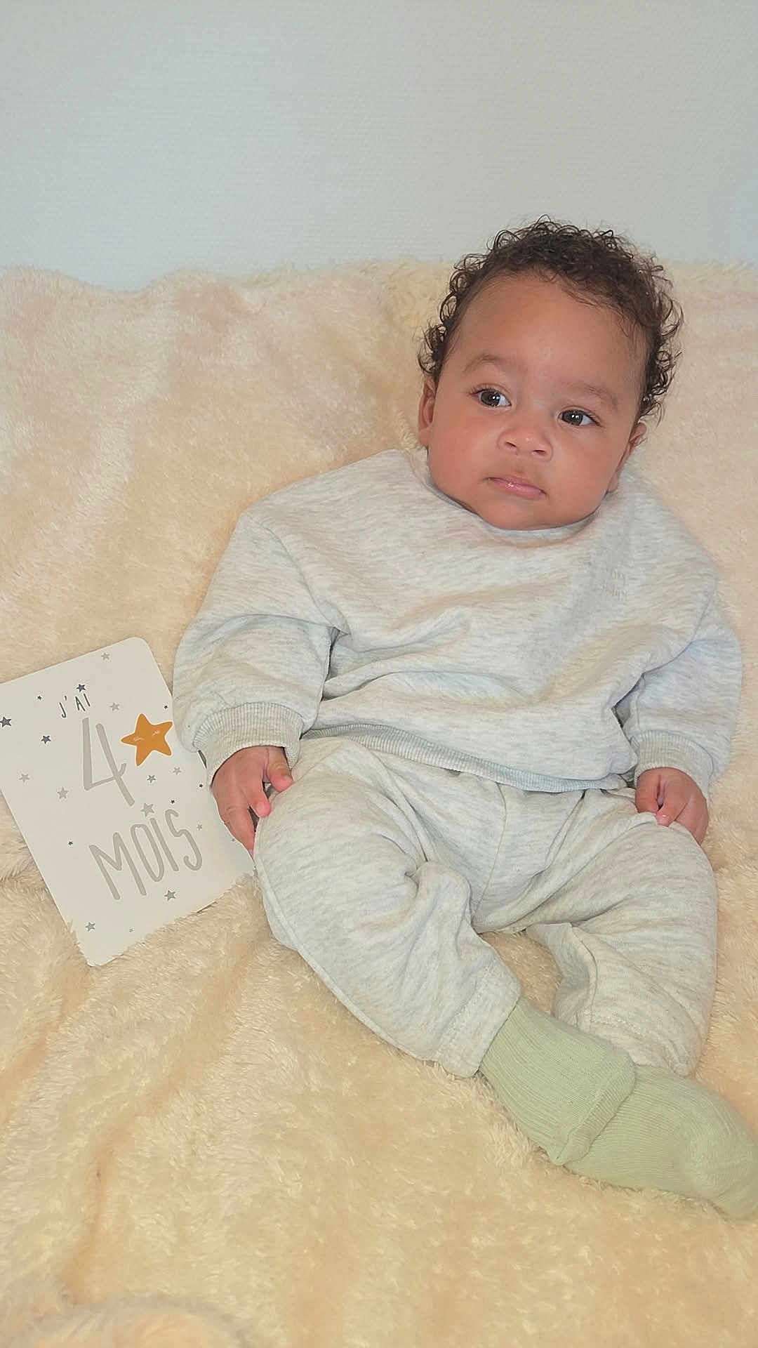 Sehyan participe au concours pour gagner de l'argent avec cette photo : baby, infant, milestone_card, gray_clothing, curly_hair, soft_blanket, socks, portrait, cute, child, indoors, sitting, milestone, celebration, skin, face, hand, cozy, young_child, resting