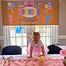 toddler, child, birthday_party, pink_tutu, table, banner, balloons, indoor, smiling, pigtails, decoration, daylight, glass_doors, celebration, happy, person, party, flower_vase, ribbon, festive