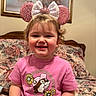 Ava is registered to the contest to win money with this photo: toddler, child, smile, pink_shirt, mouse_ears, bow, bed, floral_bedspread, painting, indoor, happy, curly_hair, portrait, cute, seated, person, headwear, decor, wall_art, casual_clothing