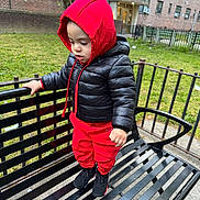 Tyler is registered to the contest to win money with this photo: air_conditioner, bench, black_jacket, building, child, daylight, fence, footwear, grass, hoodie, metal, outdoor, person, playground, red_clothing, sidewalk, standing, toddler, urban, windows