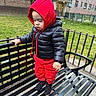 air_conditioner, bench, black_jacket, building, child, daylight, fence, footwear, grass, hoodie, metal, outdoor, person, playground, red_clothing, sidewalk, standing, toddler, urban, windows
