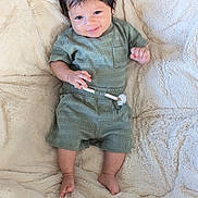 Azari is registered to the contest to win money with this photo: baby, child, infant, smile, happy, green_outfit, shorts, tshirt, dark_hair, barefoot, blanket, soft_texture, lying_down, cute, portrait, indoors, cozy, small_hands, small_feet, comfortable