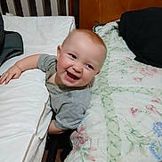 River is registered to the contest to win money with this photo: baby, smiling, laughing, bed, pillow, blanket, wooden_headboard, indoors, child, happy, cute, person, sleepwear, furniture, room, infant, expression, toddler, comfort, candid