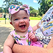 Angelique joined the competition — help win amazing prizes! baby, child, smile, laughing, pink_bow, pink_dress, outdoor, sunny, park, greenery, adult_arm, happy, headwear, person, clothing, daylight, nature, holding, toddler, portrait