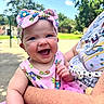 baby, child, smile, laughing, pink_bow, pink_dress, outdoor, sunny, park, greenery, adult_arm, happy, headwear, person, clothing, daylight, nature, holding, toddler, portrait