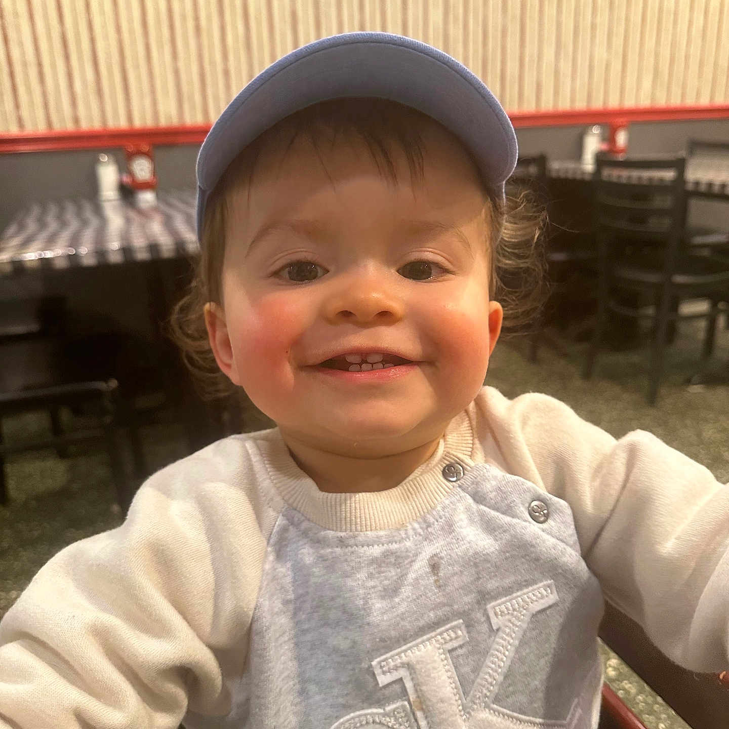 Oliver joined the competition — help win amazing prizes! toddler, child, smiling, face, cap, sweater, indoor, restaurant, table, chair, wall_art, checkered_tablecloth, hair, person, portrait, happy, cute, seated, baby, clothing
