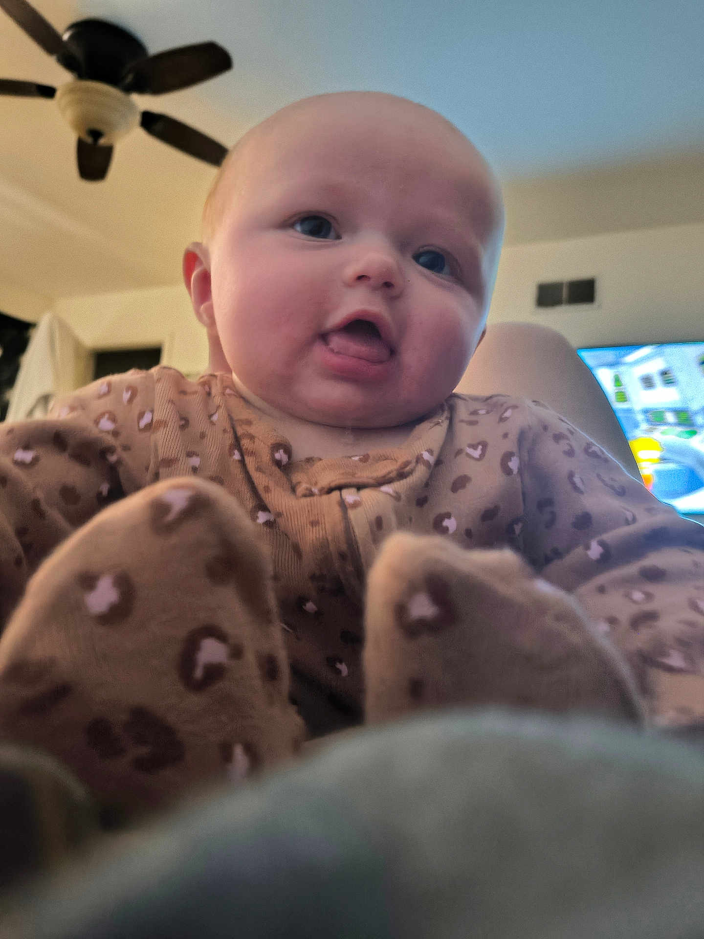 Phoebe is registered to the contest to win money with this photo: baby, infant, face, tongue, leopard_print, clothing, onesie, indoor, ceiling_fan, television, blurred_background, child, cute, playful, expression, soft_lighting, person, head, portrait, cozy