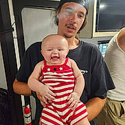 Phoebe is registered to the contest to win money with this photo: baby, child, man, smile, holding, striped_clothing, indoor, cap, black_tshirt, fire_extinguisher, person, happy, facial_expression, casual_clothing, portrait, people, arms, background, light, candid