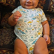 Prince Daniel is registered to the contest to win money with this photo: baby, child, smiling, dinosaur_onesie, couch, patterned_fabric, indoor, happy, sitting, cute, infant, portrait, young_child, chubby_legs, clothing, furniture, person, face, skin, hair