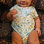 baby, child, smiling, dinosaur_onesie, couch, patterned_fabric, indoor, happy, sitting, cute, infant, portrait, young_child, chubby_legs, clothing, furniture, person, face, skin, hair