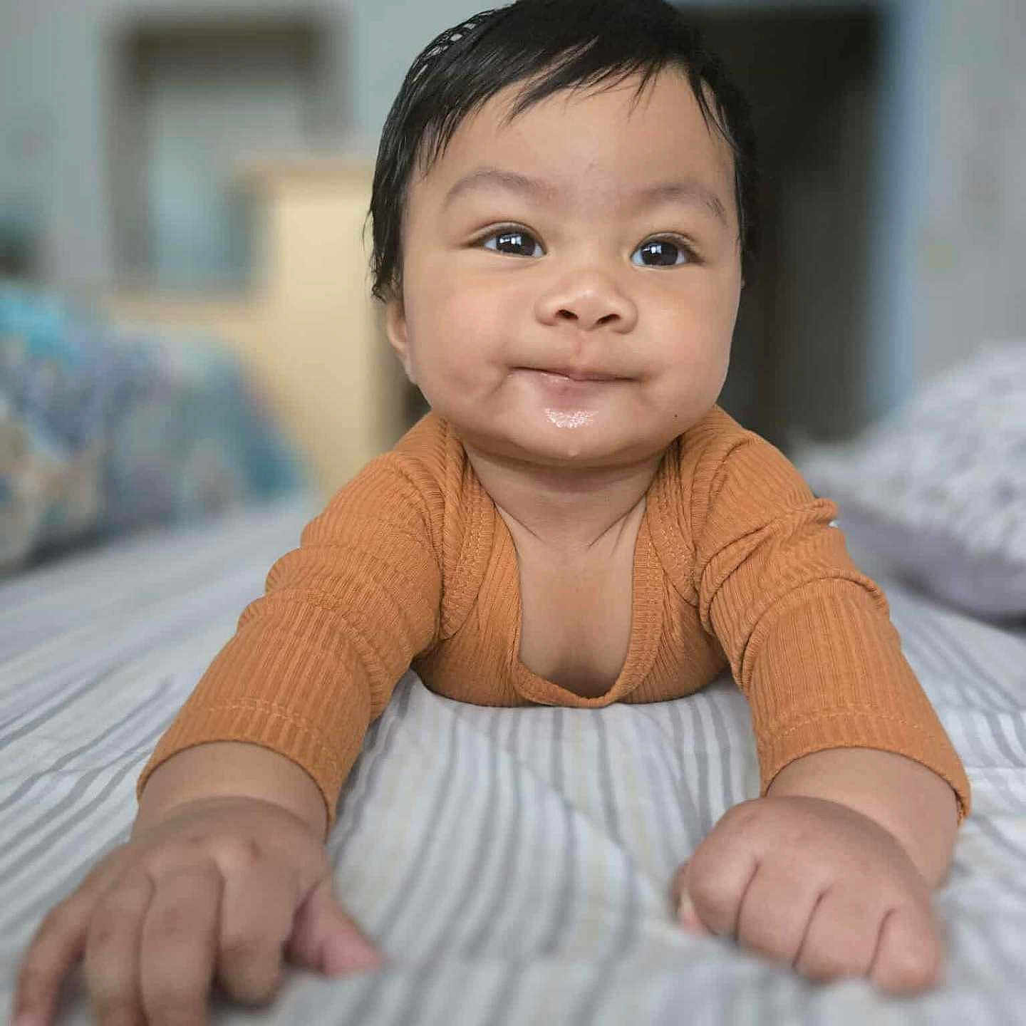 Prince Daniel is registered to the contest to win money with this photo: baby, bed, blurred_background, child, closeup, cozy, cute, face, hands, happy, indoor, infant, lying_down, orange_clothing, person, portrait, skin, smile, striped_bedspread, young_child