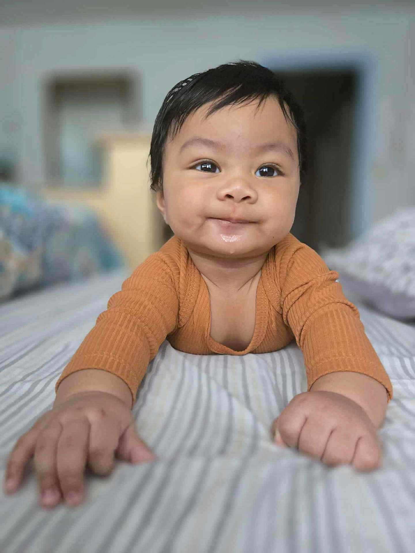 Prince Daniel is registered to the contest to win money with this photo: baby, child, infant, smile, face, hands, orange_clothing, bed, striped_bedspread, indoor, person, cute, portrait, lying_down, skin, young_child, closeup, happy, blurred_background, cozy