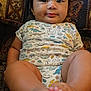Prince Daniel is registered to the contest to win money with this photo: baby, infant, child, onesie, dinosaur_print, feet, legs, face, hair, couch, patterned_fabric, comfort, indoors, portrait, cute, smiling, skin, relaxed, seated, closeup