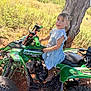 child, girl, atv, green_vehicle, outdoor, tree, grass, boots, denim_dress, sunlight, nature, dirt, young_person, vehicle, portrait, blue_eyes, curly_hair, playful, adventure, rural