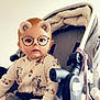 baby, child, stroller, onesie, patterned_clothing, face_filter, bear_ears, glasses_filter, indoor, infant, cute, seated, hand, expression, light_background, soft_lighting, toy, blanket, person, portrait