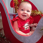 River-Lee is registered to the contest to win money with this photo: baby, child, red_clothing, sleigh, holiday_decor, gold_tinsel, ornament, smile, face, indoors, festive, cute, happy, decorative, furniture, christmas_theme, cheerful, person, seat, celebration