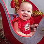 baby, child, red_clothing, sleigh, holiday_decor, gold_tinsel, ornament, smile, face, indoors, festive, cute, happy, decorative, furniture, christmas_theme, cheerful, person, seat, celebration