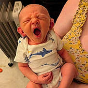 Josiah is registered to the contest to win money with this photo: baby, infant, yawning, onesie, shark_pattern, holding, arm, person, indoor, heater, yellow_clothing, floral_pattern, candid, cute, newborn, foot, hand, expression, casual, skin