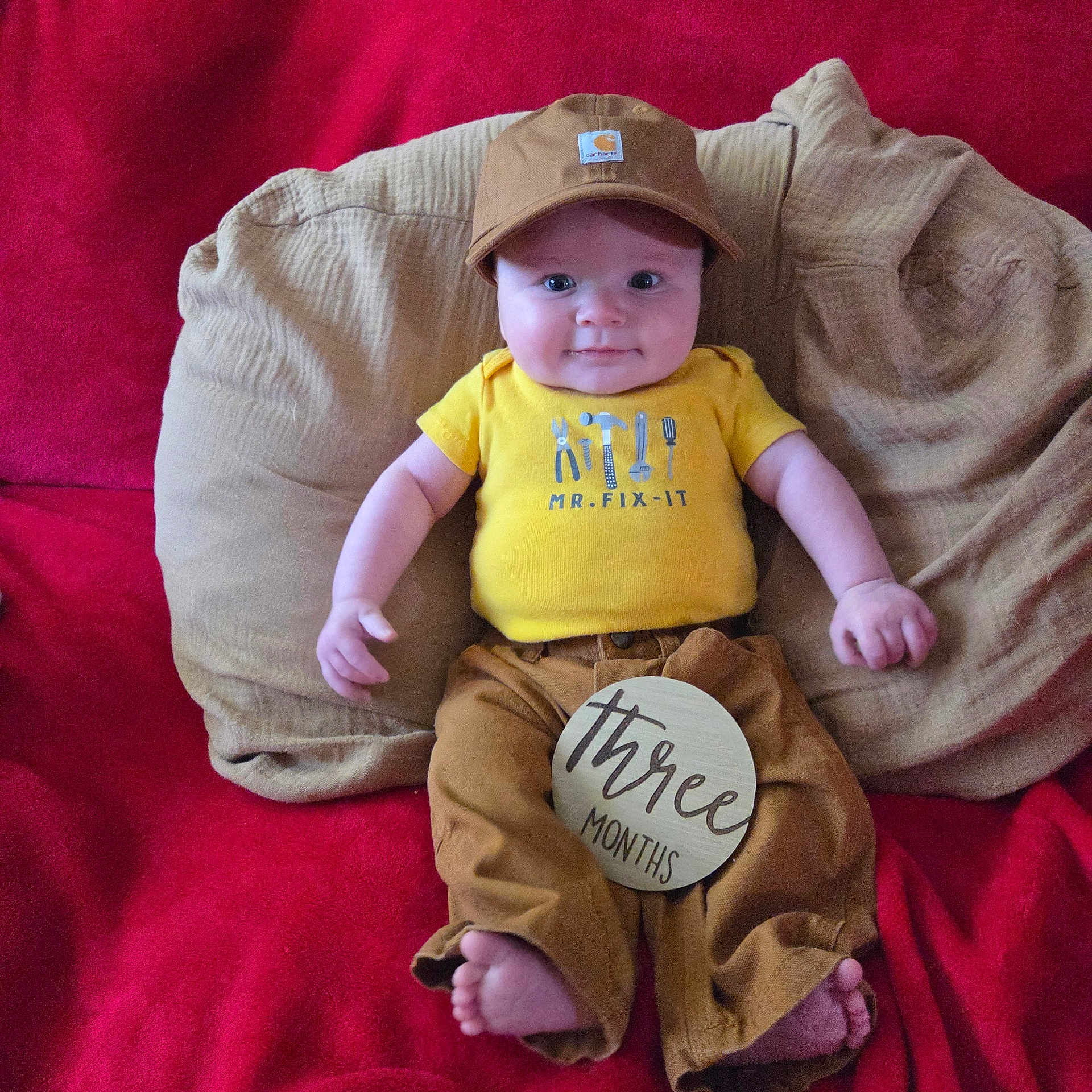 Waylon joined the competition — help win amazing prizes! baby, infant, child, cap, hat, yellow_shirt, brown_pants, cushion, red_blanket, milestone_sign, three_months, smiling, cute, portrait, indoors, person, sitting, clothing, happy, face