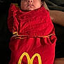 baby, sleeping, blanket, red, yellow, logo, mcdonalds, child, swaddled, infant, face, closed_eyes, person, resting, cute, indoors, fabric, hand, soft, cozy