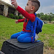 Zion Wesley is registered to the contest to win money with this photo: child, toddler, costume, spiderman, backpack, outdoor, grass, sky, cloudy, utility_box, stone_building, greenery, side_view, playful, cute, small_child, sitting, reaching, casual, daylight