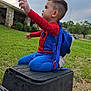 child, toddler, costume, spiderman, backpack, outdoor, grass, sky, cloudy, utility_box, stone_building, greenery, side_view, playful, cute, small_child, sitting, reaching, casual, daylight