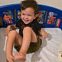 child, smiling, bed, plush_toy, paw_patrol, headboard, black_shirt, shorts, indoors, happy, playful, toy, furniture, wall, foot, person, cute, laughing, young_child, room