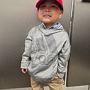 Seth is registered to the contest to win money with this photo: child, boy, red_cap, mario_hat, grey_hoodie, adidas, beige_pants, standing, smiling, playful, indoors, metallic_background, door, casual_clothing, portrait, person, cute, expression, young, confident