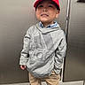 child, boy, red_cap, mario_hat, grey_hoodie, adidas, beige_pants, standing, smiling, playful, indoors, metallic_background, door, casual_clothing, portrait, person, cute, expression, young, confident