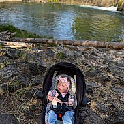 Octavia is registered to the contest to win money with this photo: baby, car_seat, infant, pink_bow, jeans, socks, rocky_ground, river, water, trees, nature, outdoor, sunlight, grass, seatbelt, child, headband, person, scenic, relaxing