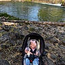 baby, car_seat, infant, pink_bow, jeans, socks, rocky_ground, river, water, trees, nature, outdoor, sunlight, grass, seatbelt, child, headband, person, scenic, relaxing