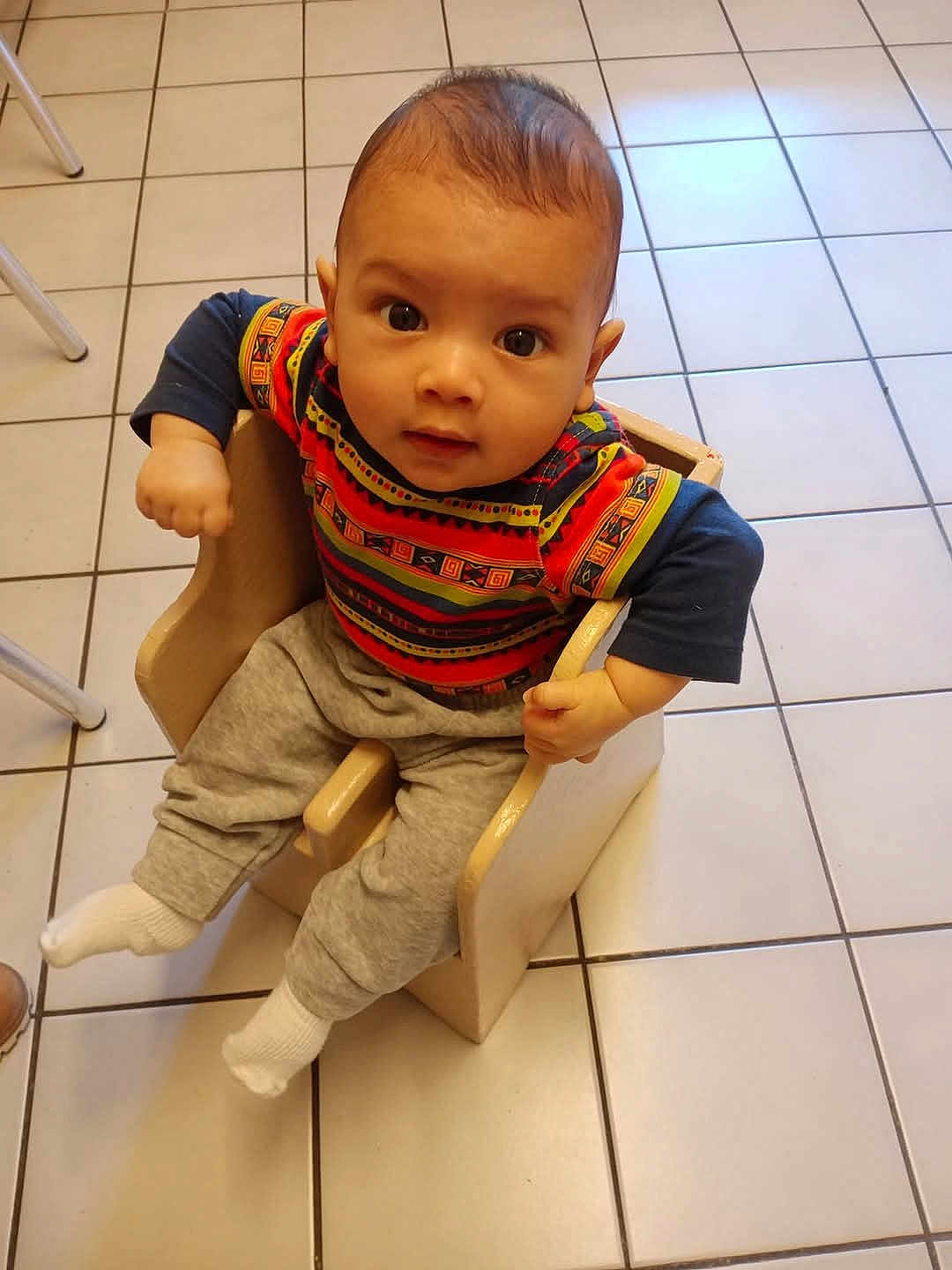 Léo participe au concours pour gagner de l'argent avec cette photo : baby, child, toddler, chair, colorful_clothing, patterned_shirt, gray_pants, white_socks, tiled_floor, indoor, cute, looking_up, wooden_chair, seated, small_hands, curious, floor_tiles, young_child, casual_wear, infant