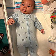 Alexander is registered to the contest to win money with this photo: baby, infant, dinosaur_onesie, changing_pad, baby_toy, baby_care_items, footwear, striped_slides, face, portrait, lying_down, baby_bottle, skincare_products, blue_clothing, cute, indoor, newborn, baby_changing_station, expression, hand